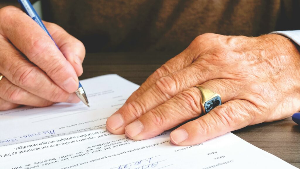 "Manos de una persona mayor firmando un documento con bolígrafo azul sobre una mesa, usando un anillo dorado con escudo grabado. Leyendo el decreto 102/2025