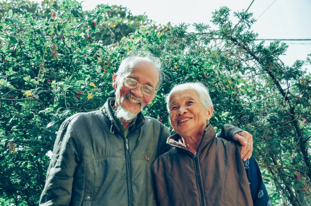 Pareja de adultos mayores sonriendo y abrazados en un jardín lleno de vegetación.