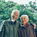 Pareja de adultos mayores sonriendo y abrazados en un jardín lleno de vegetación.